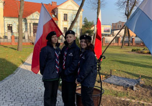 Troje harcerzy stoi przy flagach Polski.